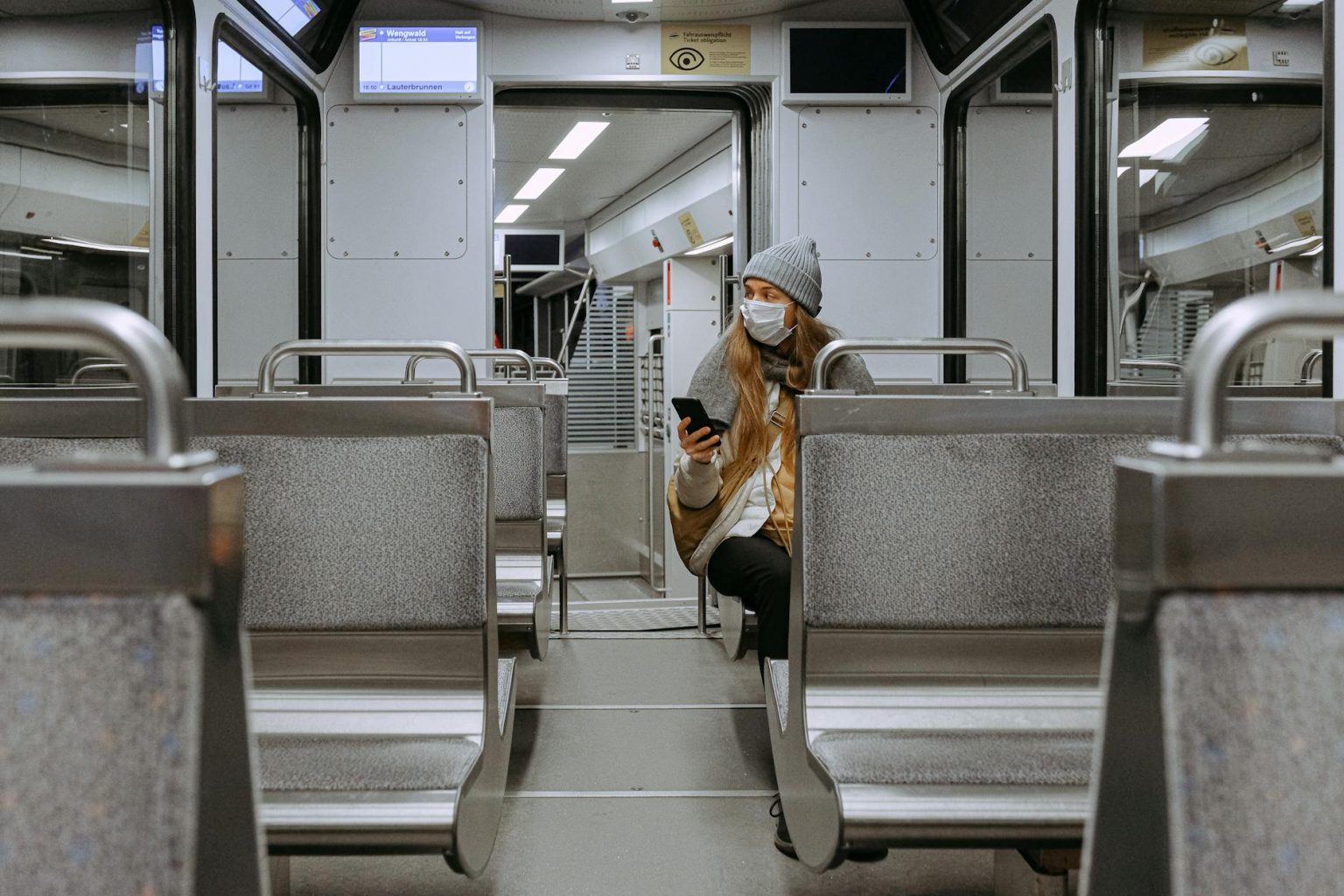 A woman wearing a face mask is sitting alone on a nearly empty urban train, practicing safety measures.