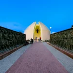 A view of a monument in Kazakhstan with sculptured walls and people at dusk. Captures cultural essence.