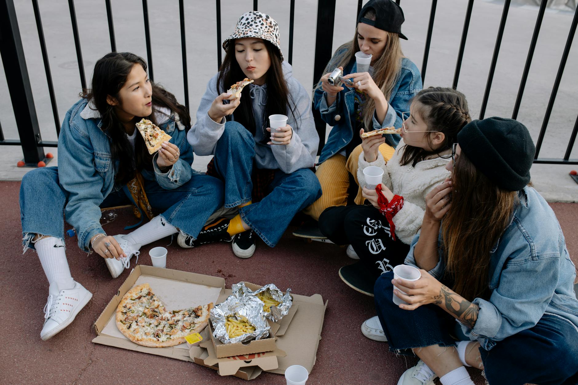 Five young women sharing pizza and drinks outdoors in a casual urban setting.