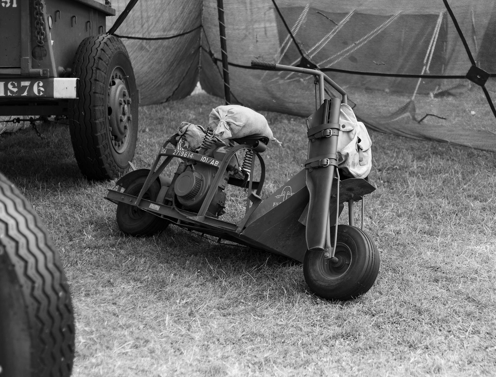 A black and white image of a vintage military scooter under a tent at a camp.