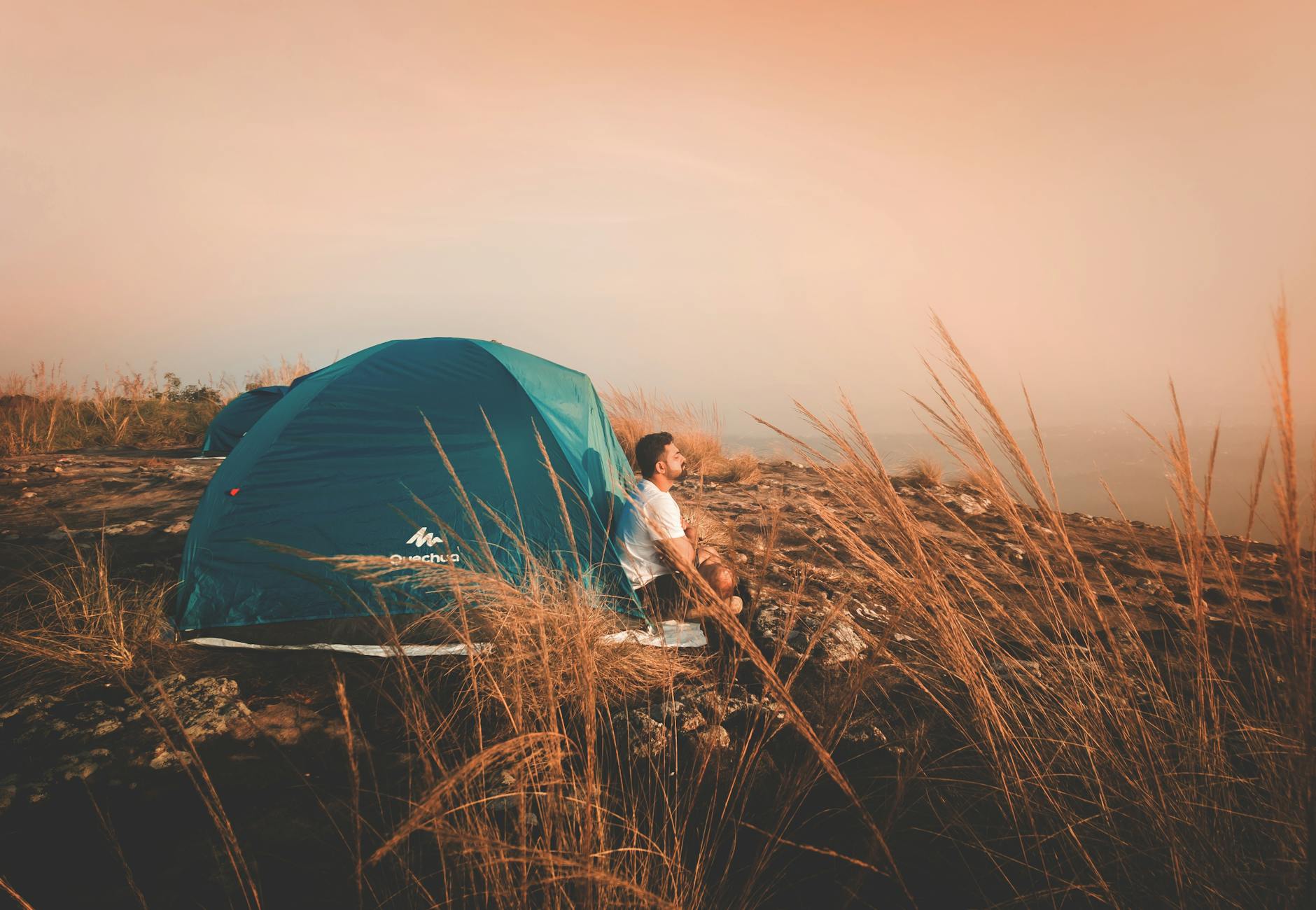 A man enjoys a peaceful camping experience beside a blue tent in a tranquil natural setting during sunset.