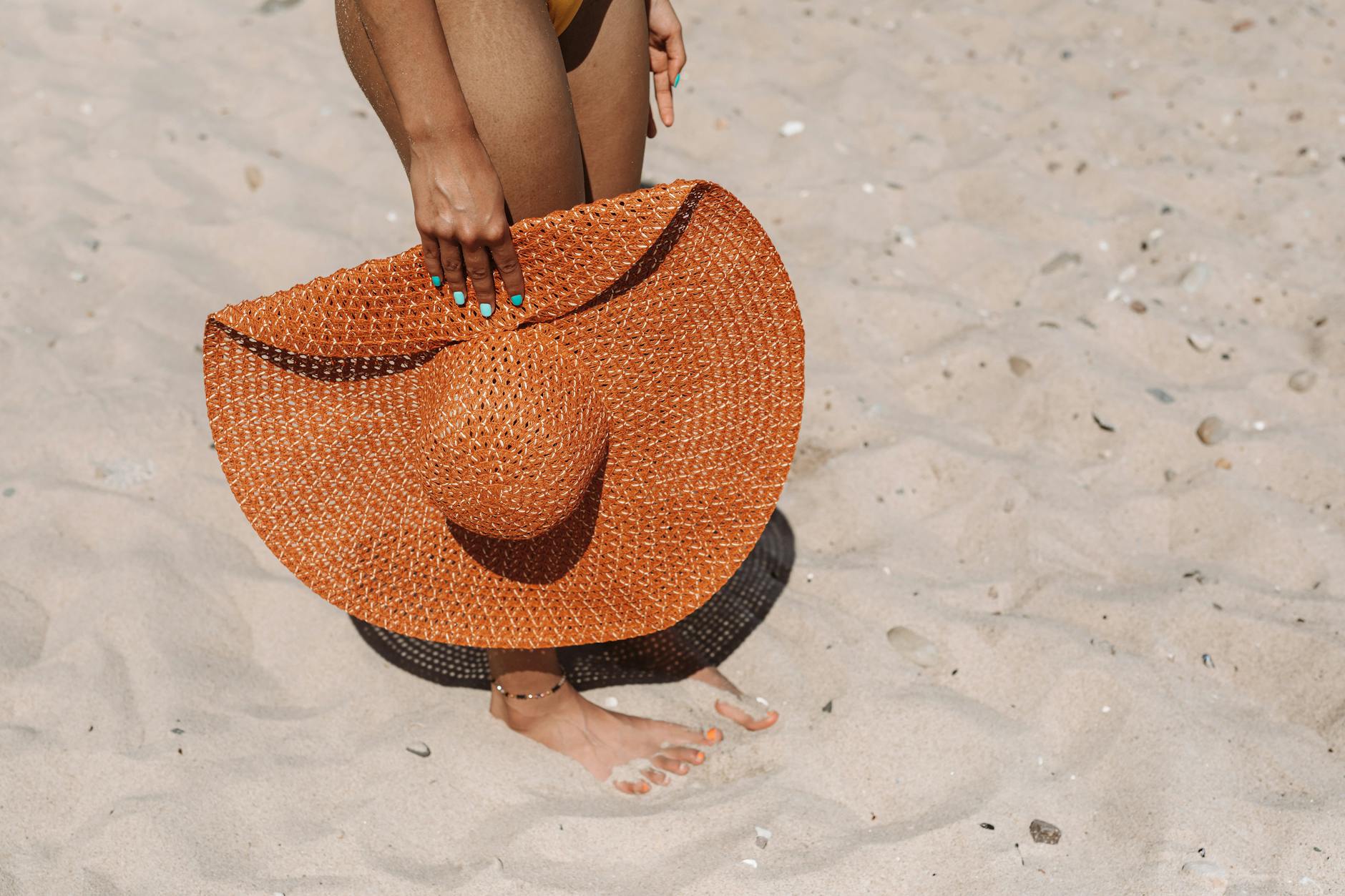 A close-up of a woman on sandy beach holding a large orange woven sunhat.