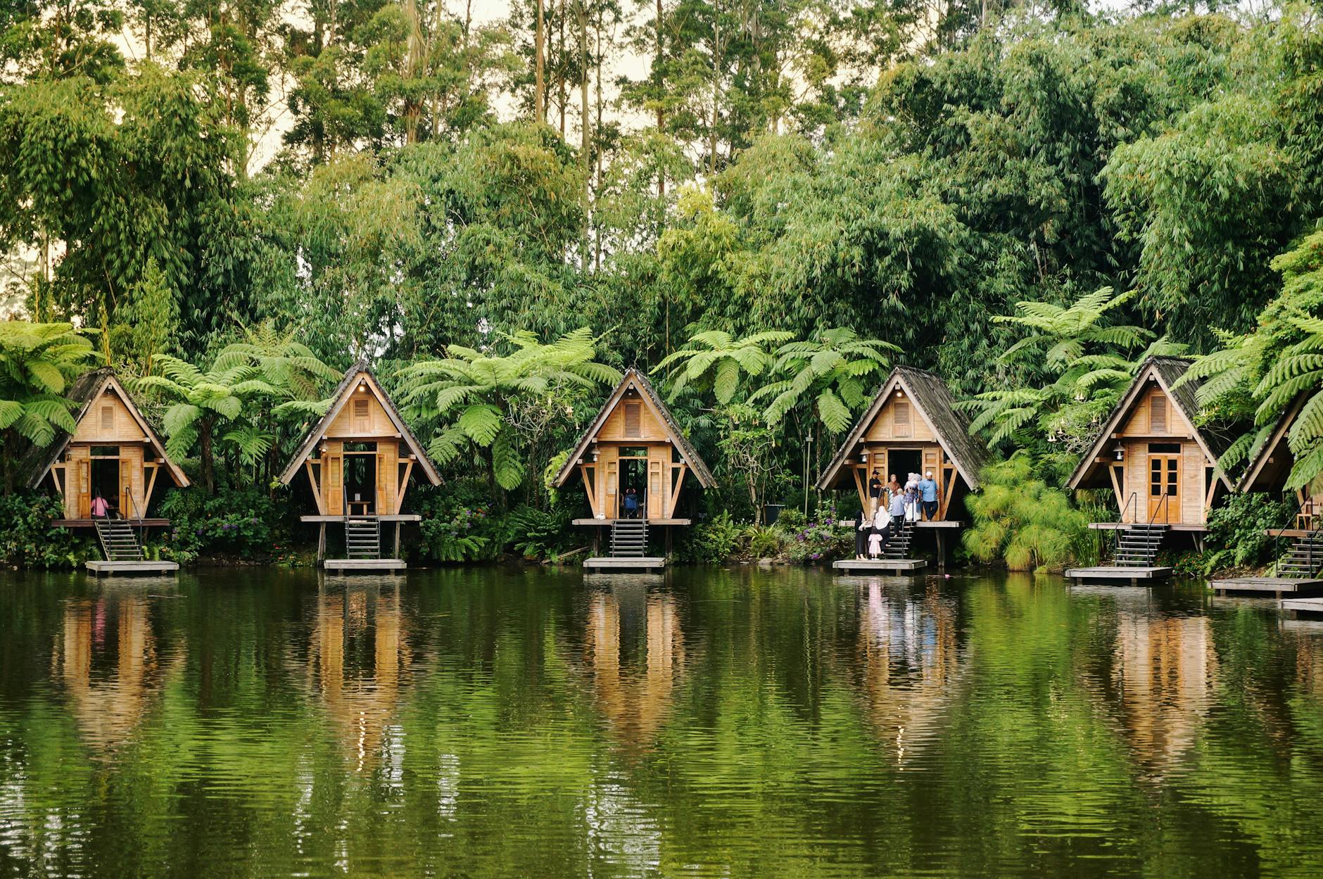 Picturesque wooden cabins reflect over a tranquil lake against lush greenery.