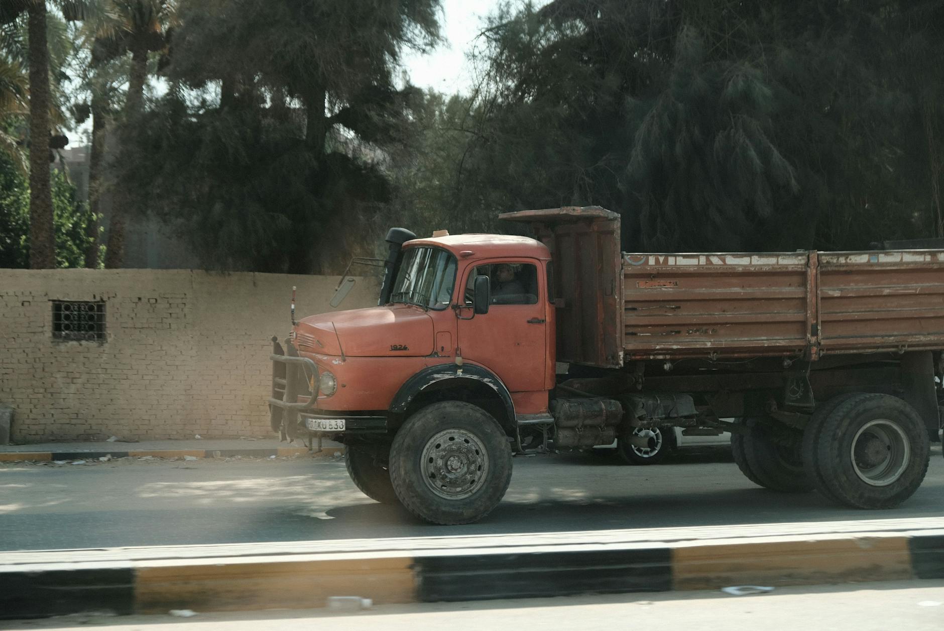 A vintage red Mercedes truck navigates a sunlit Cairo street with palm trees in the background.