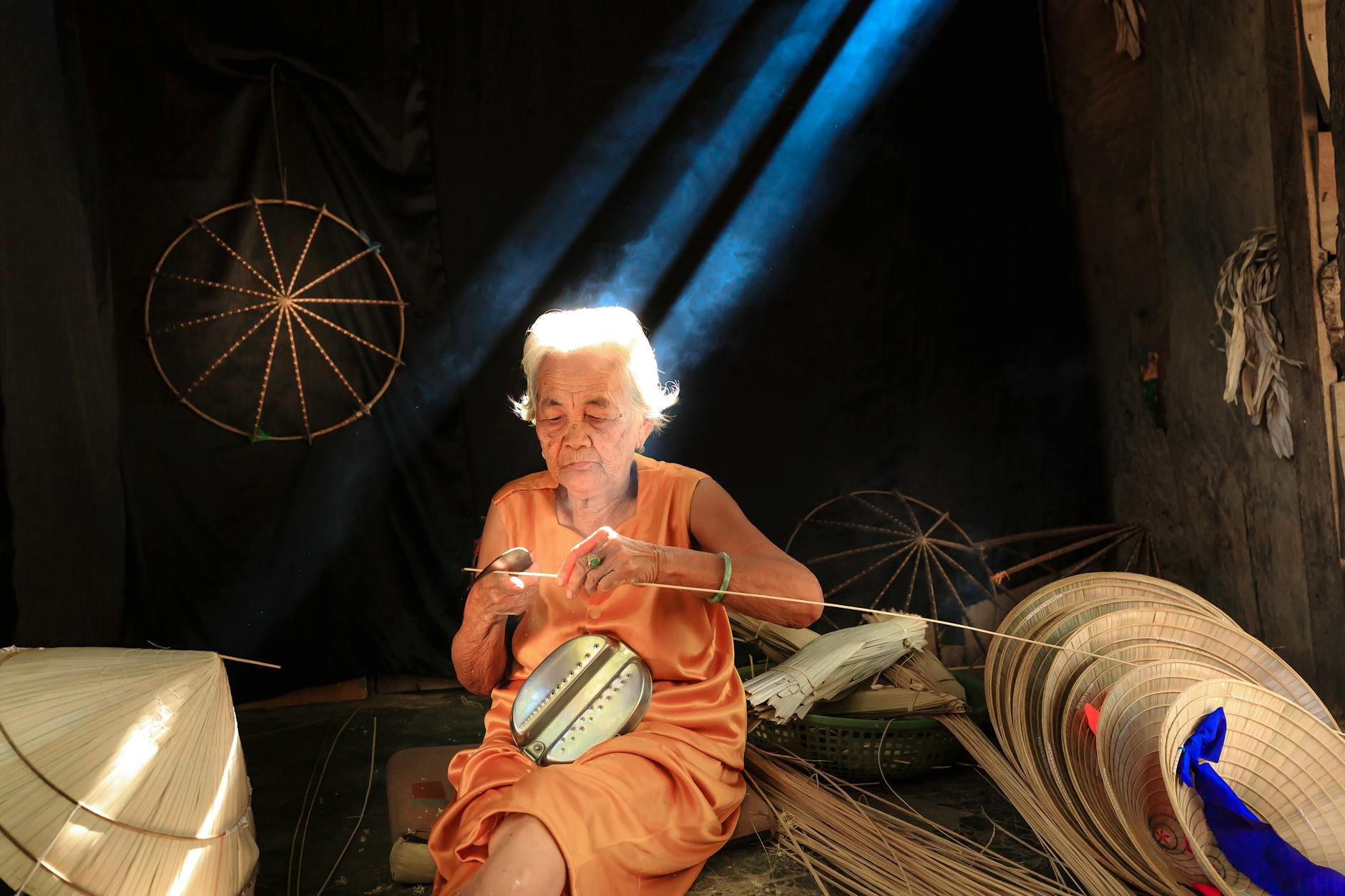 Senior woman crafting traditional hats in serene indoor setting with dramatic lighting.