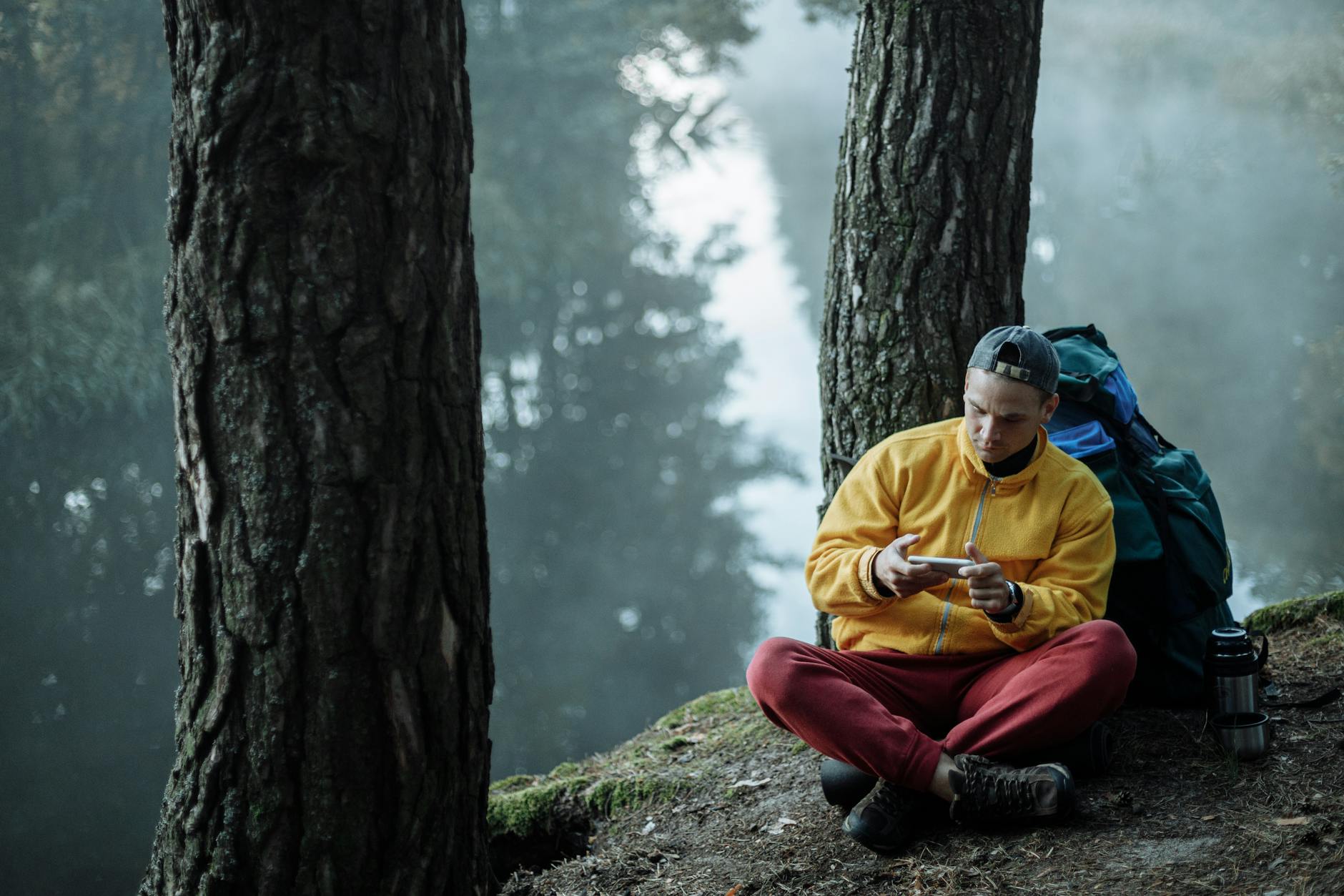 A man in a yellow jacket sits by a forest river, using a smartphone, surrounded by nature.