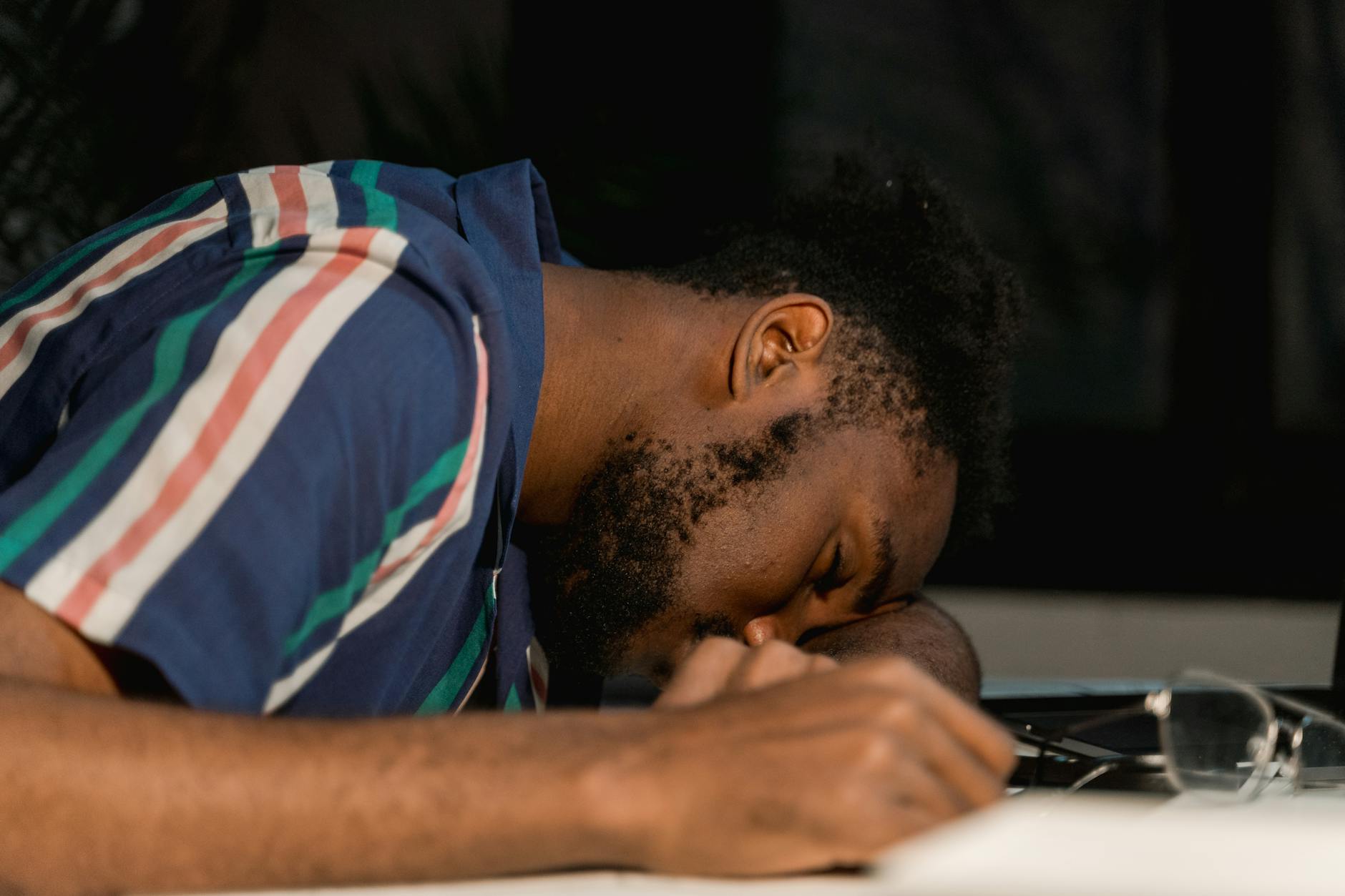A man with facial hair rests his head on a desk, napping during a late work session.