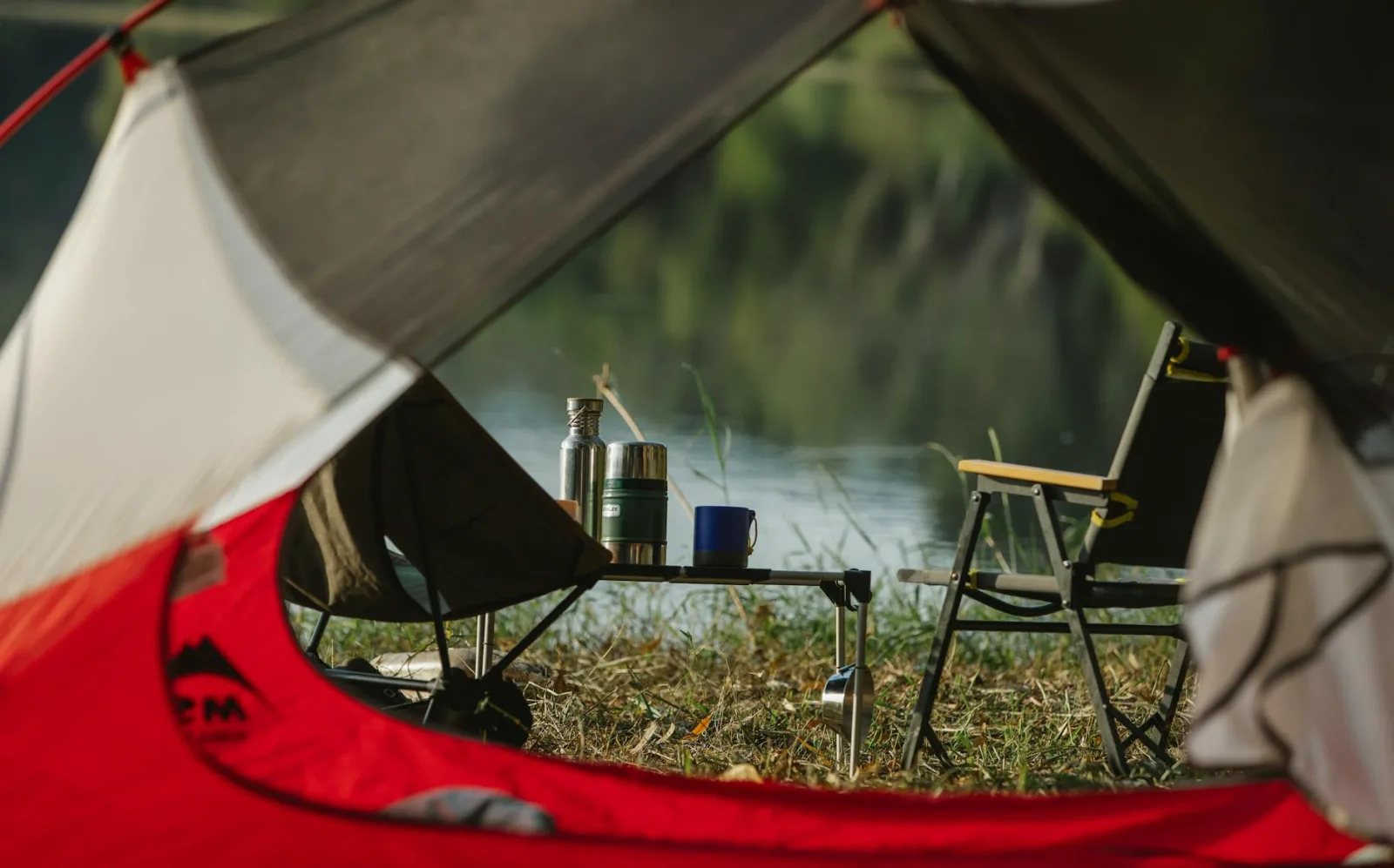Through tent hole view of portable chairs and table with thermoses on grass shore against lake