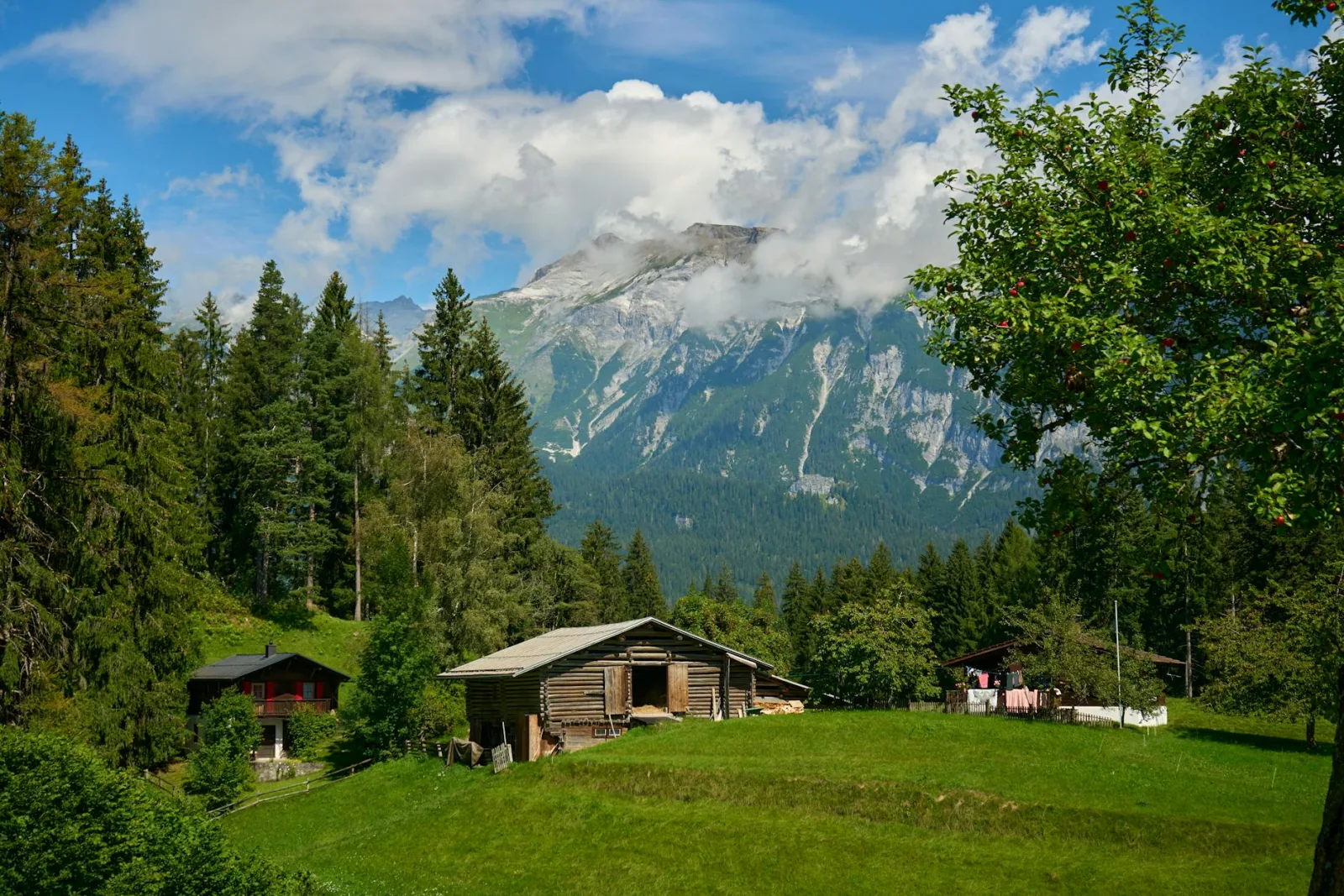 Scenic mountainous landscape with wooden cabins, trees, and lush greenery under a bright cloudy sky.