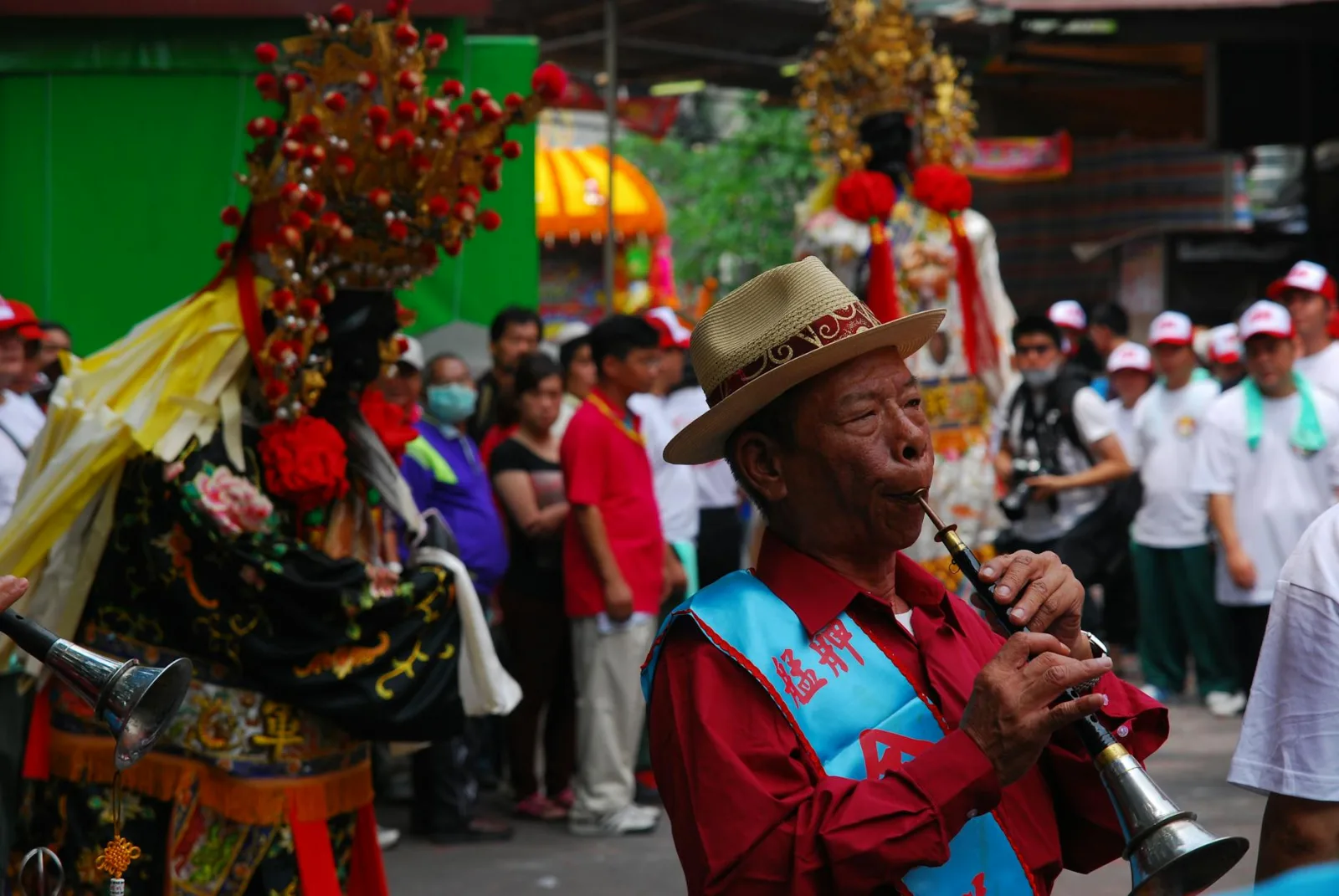 Elderly man playing traditional instrument during a cultural parade with colorful costumes.