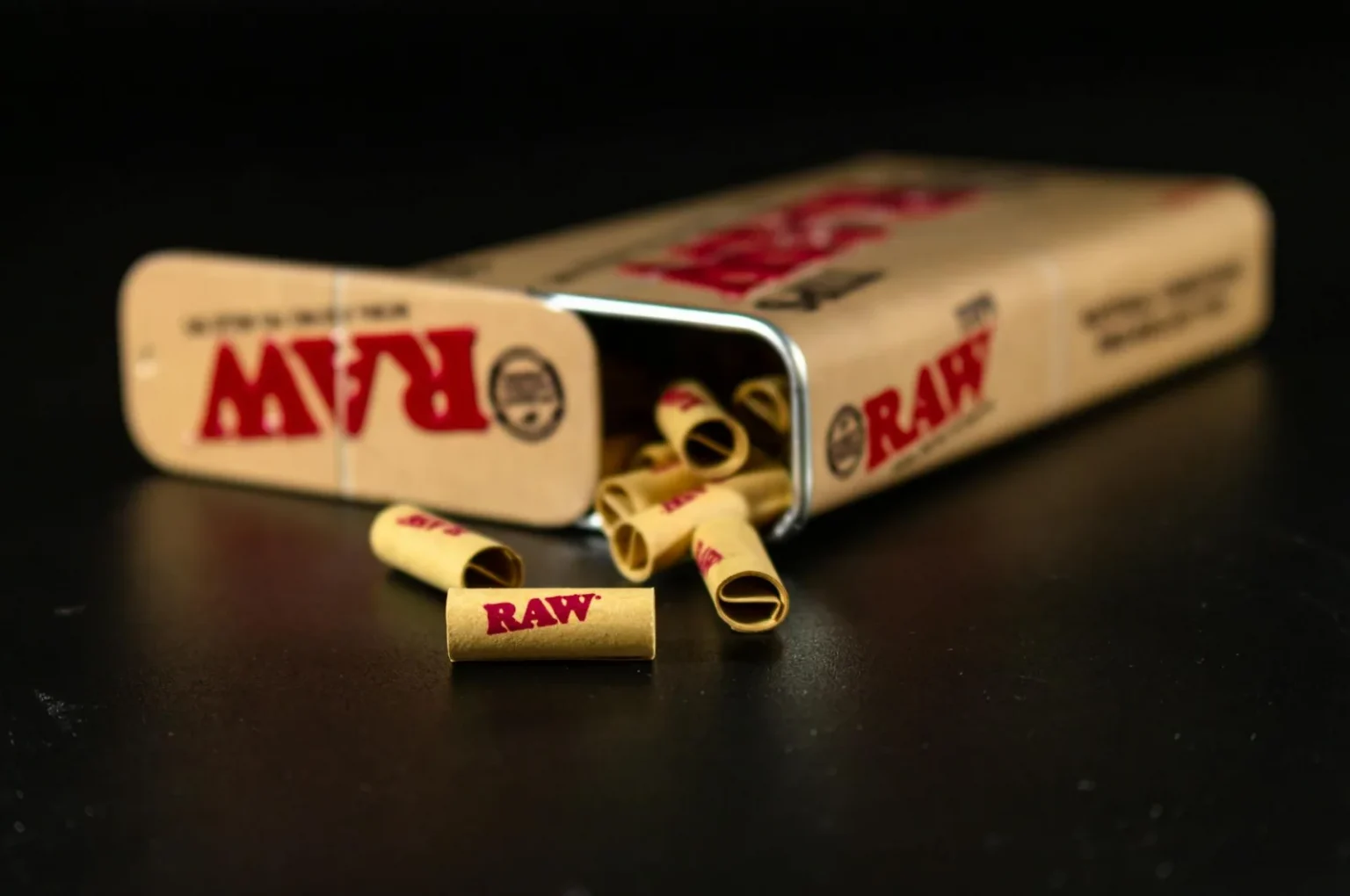 Detailed image of Raw rolling paper tips scattered from a tin box on a dark surface.
