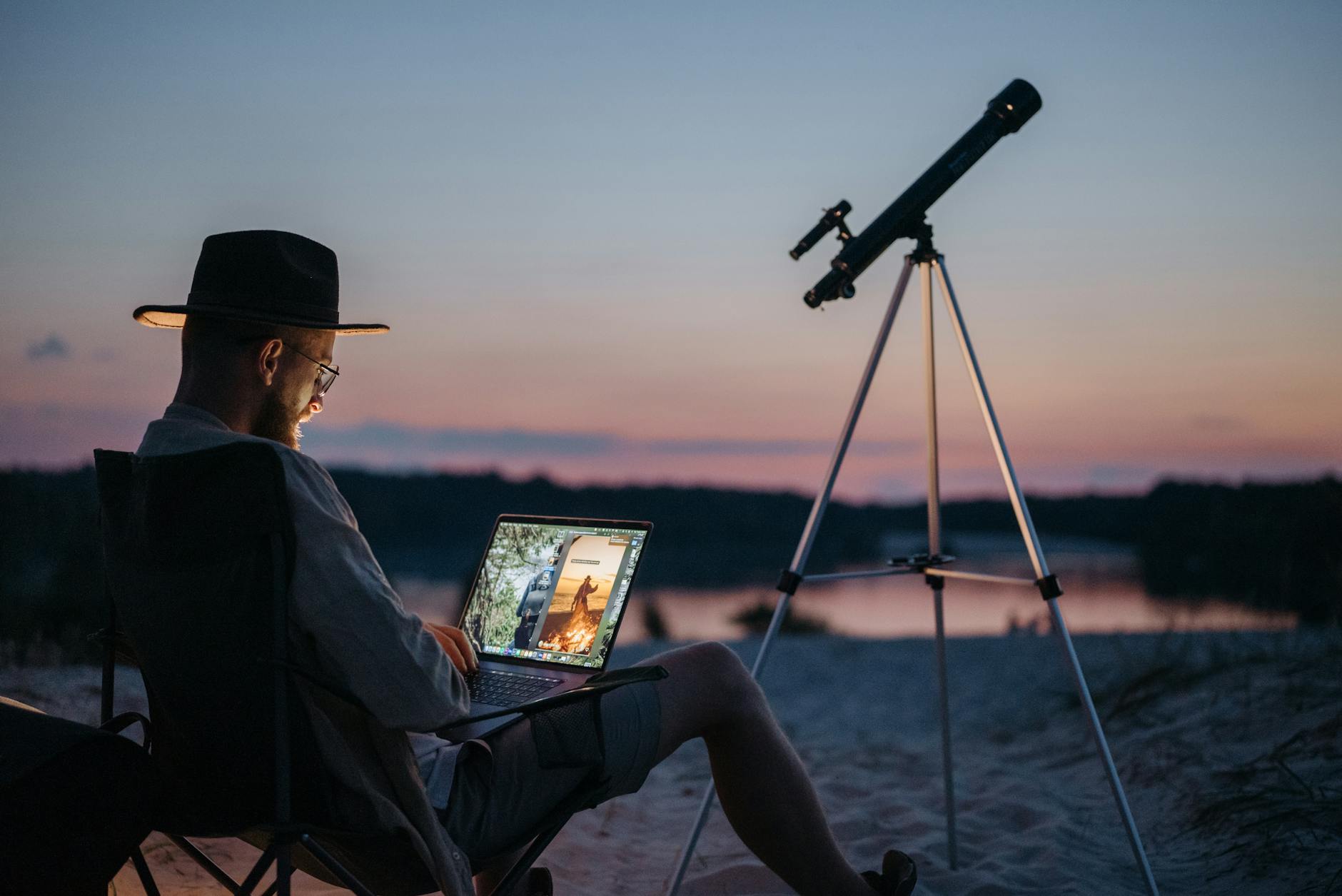 A man enjoys a camping trip by the lake using a laptop and telescope at sunset.