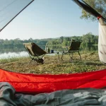 Relaxing campsite view from a tent by a peaceful lake with chairs under clear skies.