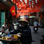 A vendor in a conical hat pushes a cart through a colorful street with red lanterns and flags.