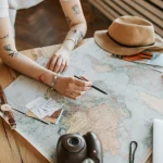 Tattooed woman planning a trip with a world map, passports, and travel essentials on a wooden table.