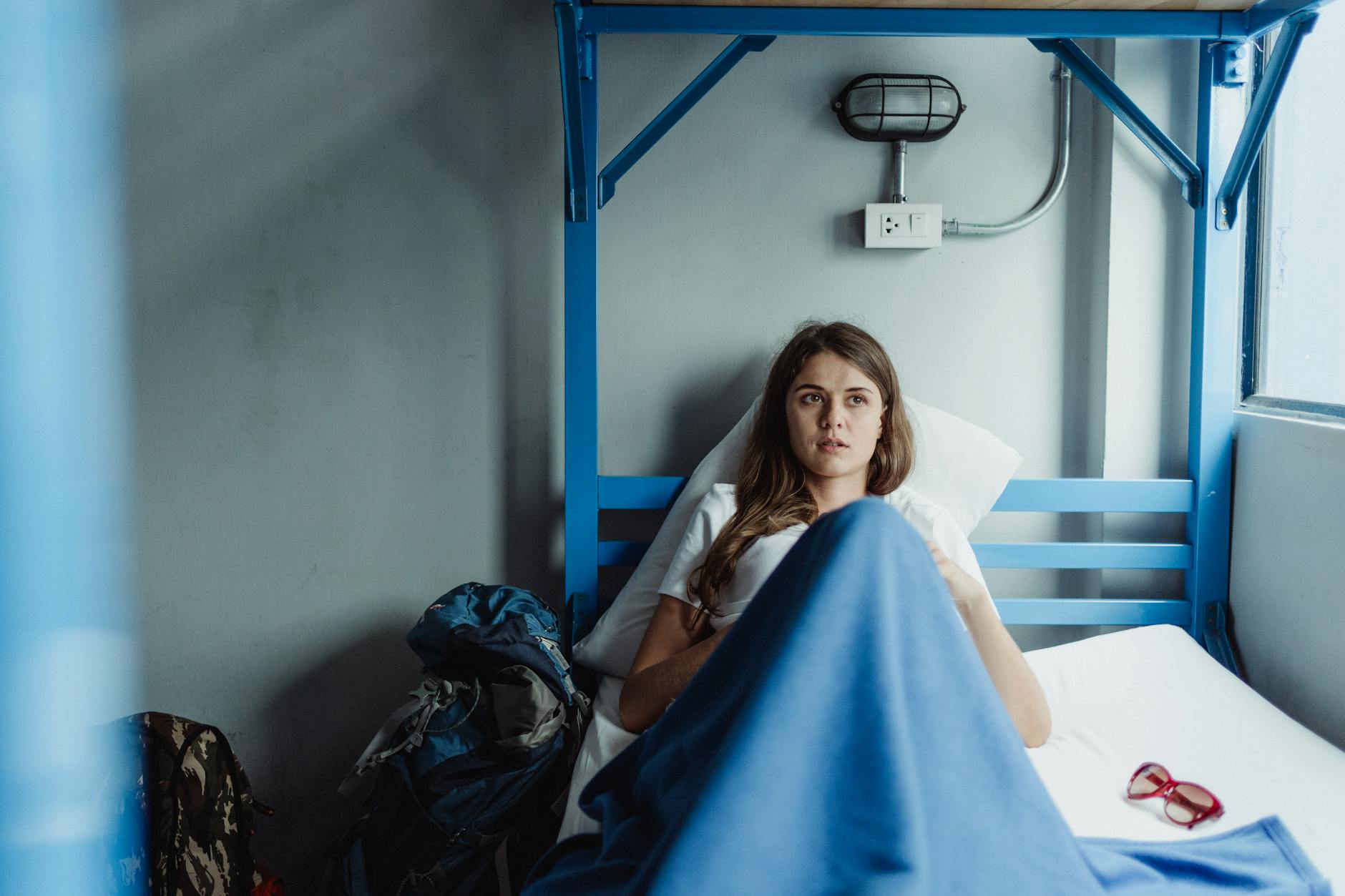 A woman lying in a hostel bed, surrounded by a backpack and glasses, enjoying relaxation.