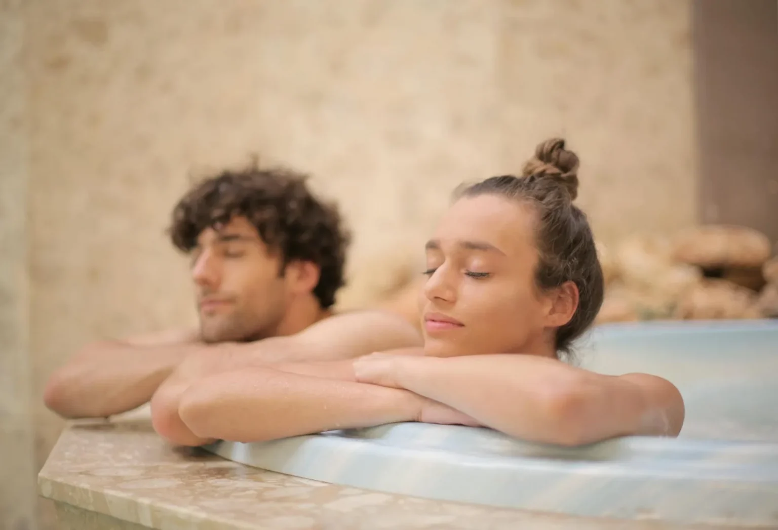 Happy cheerful man and woman with closed eyes bathing together leaning on bath edge during romantic weekend in luxury hotel in bathroom with marble interior