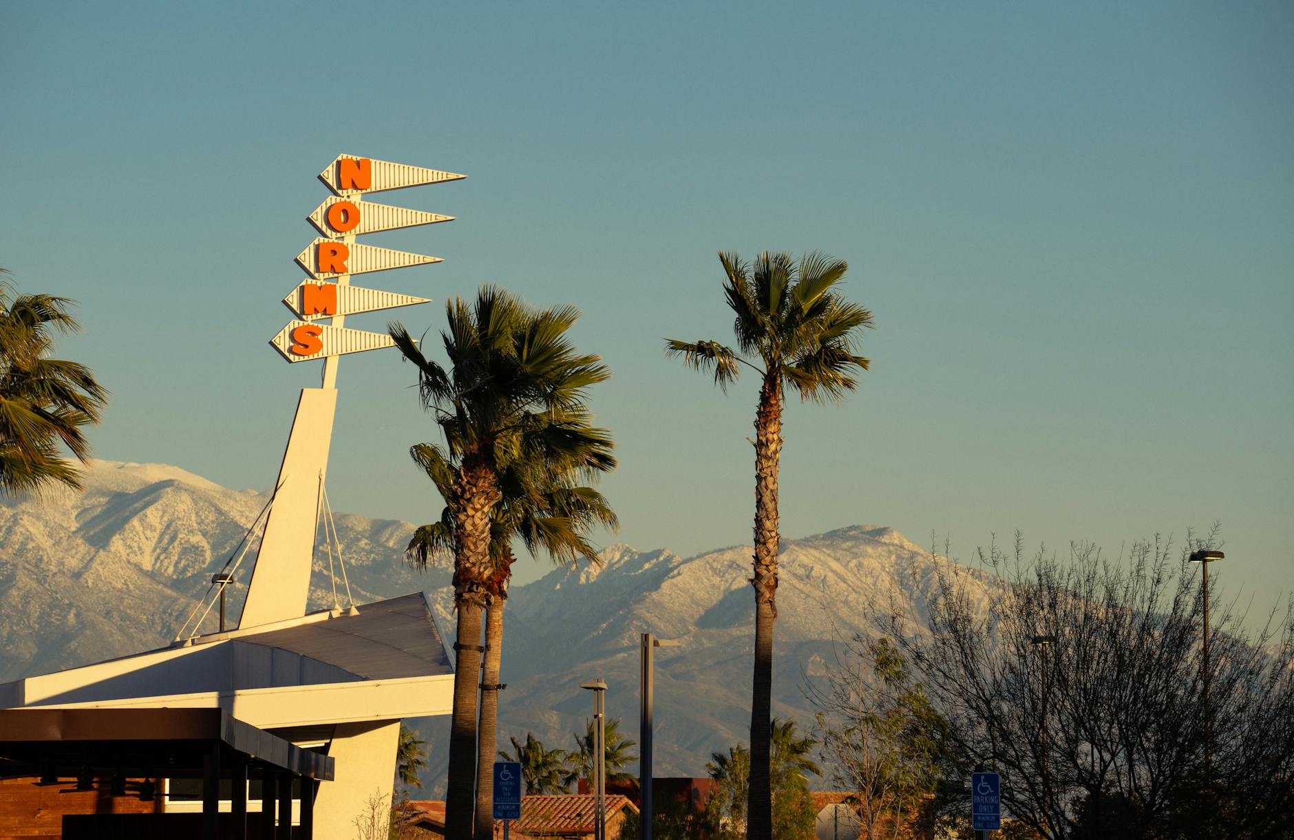 A retro Norms restaurant sign framed by palm trees and snow-capped mountains in Rialto, CA.