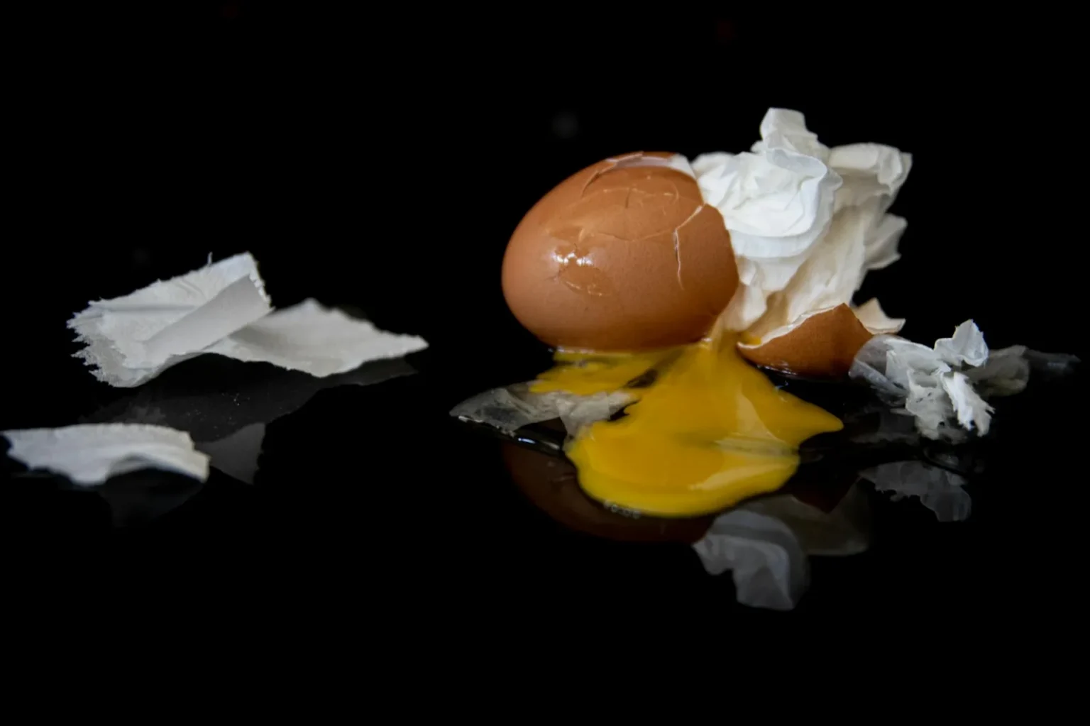 A cracked egg with yolk spread on a reflective dark surface surrounded by crumpled paper.