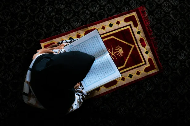 An overhead view of a woman reading the Quran on a prayer mat indoors.