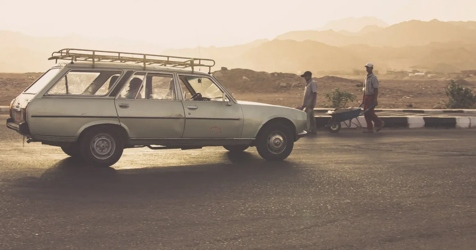 A classic Peugeot car with people walking by in Egypt's scenic desert sunset.