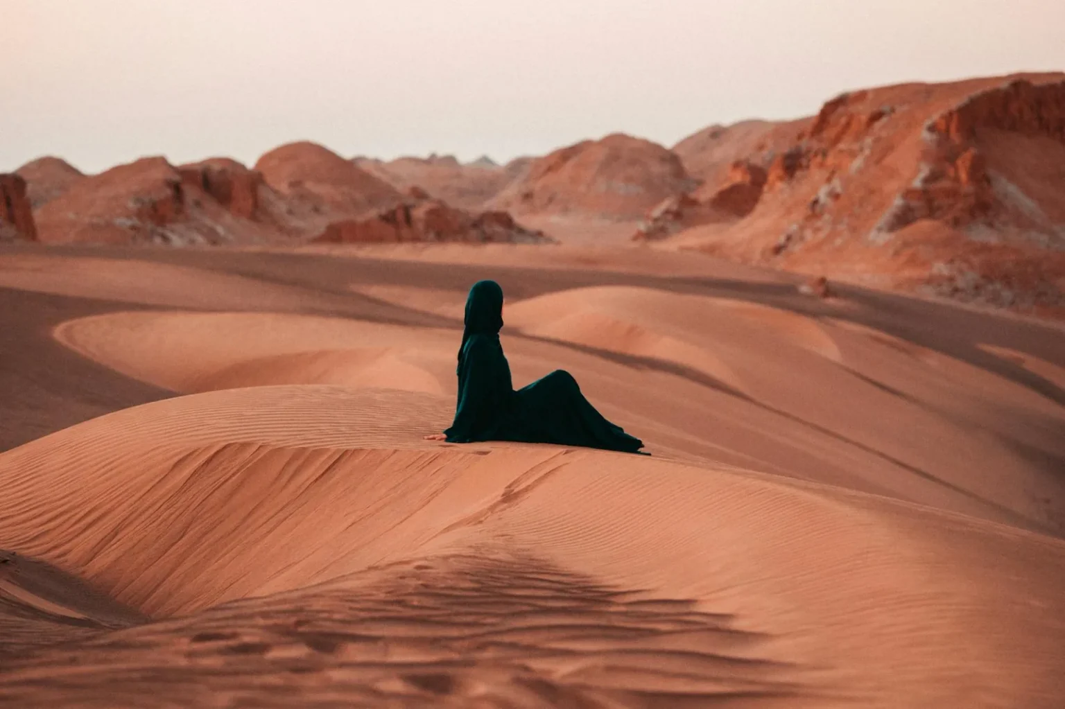 A person in traditional attire sits on a dune at sunset, embracing solitude and tranquility.
