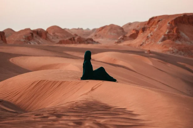 A person in traditional attire sits on a dune at sunset, embracing solitude and tranquility.