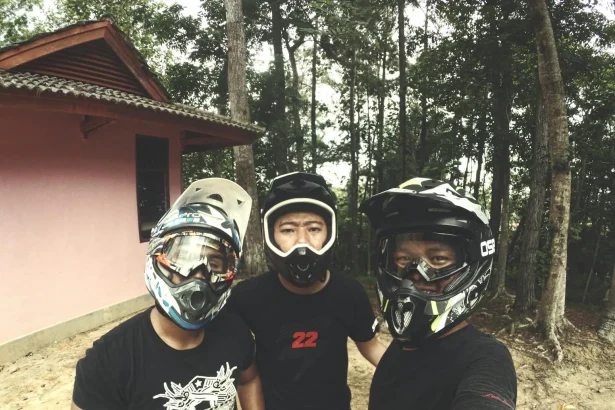Three friends in helmets taking a selfie during a dirt biking adventure in a forest setting.