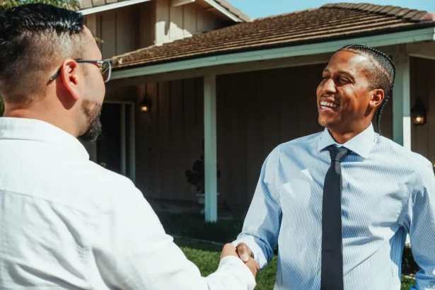 Two real estate professionals shaking hands in front of a new home during a transaction.