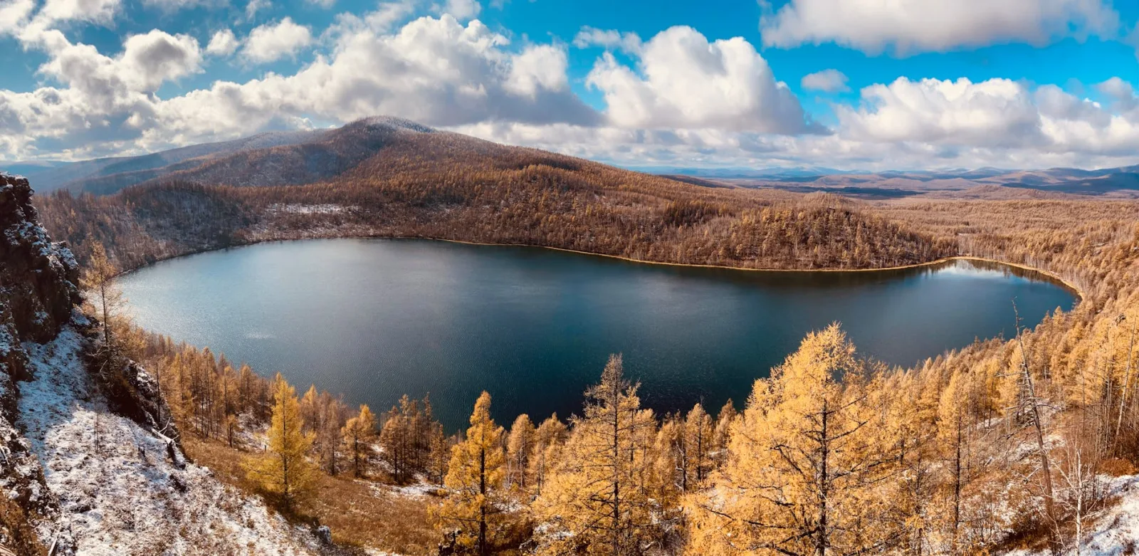 Tranquil lake surrounded by autumn forests and mountains under a bright blue sky.