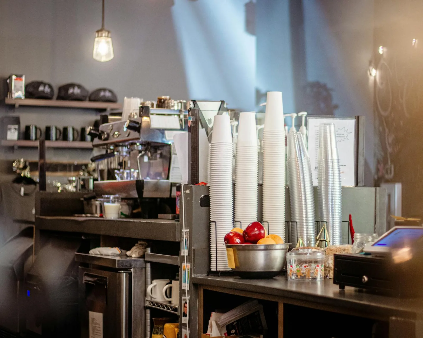 Warm café interior featuring a counter with cups, a bowl of fruit, and an espresso machine.