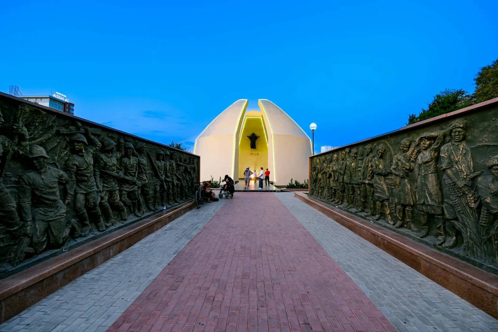 A view of a monument in Kazakhstan with sculptured walls and people at dusk. Captures cultural essence.