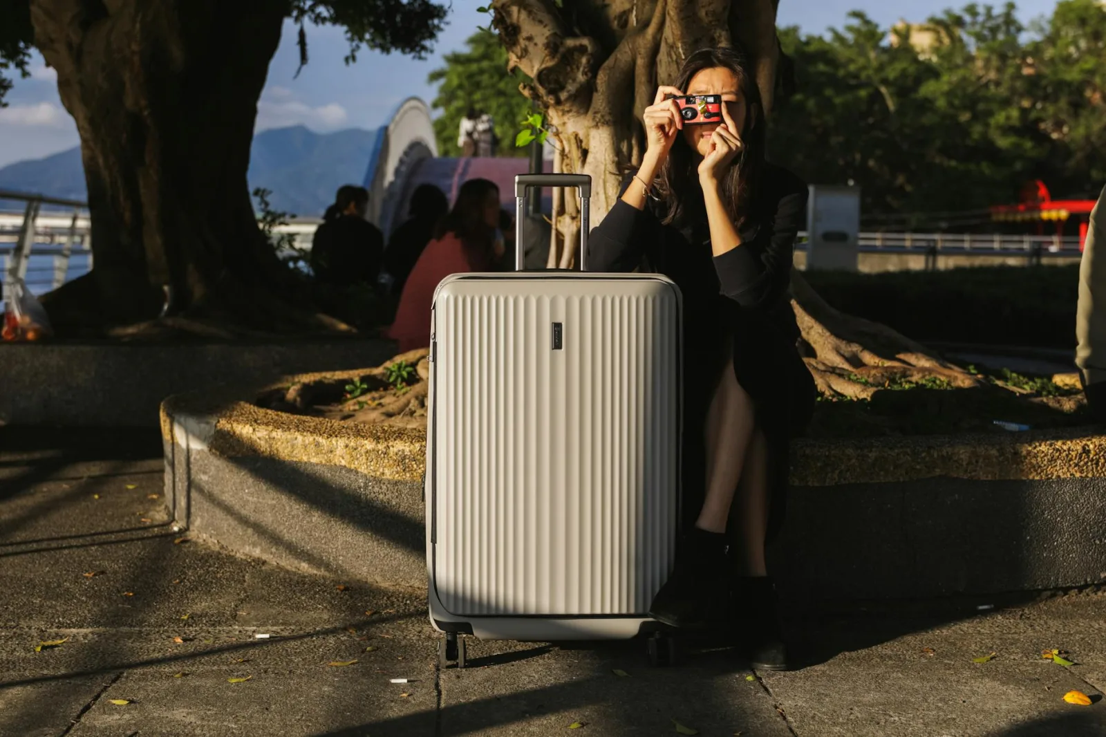 A woman with a suitcase and vintage camera enjoys photography in an urban park setting.