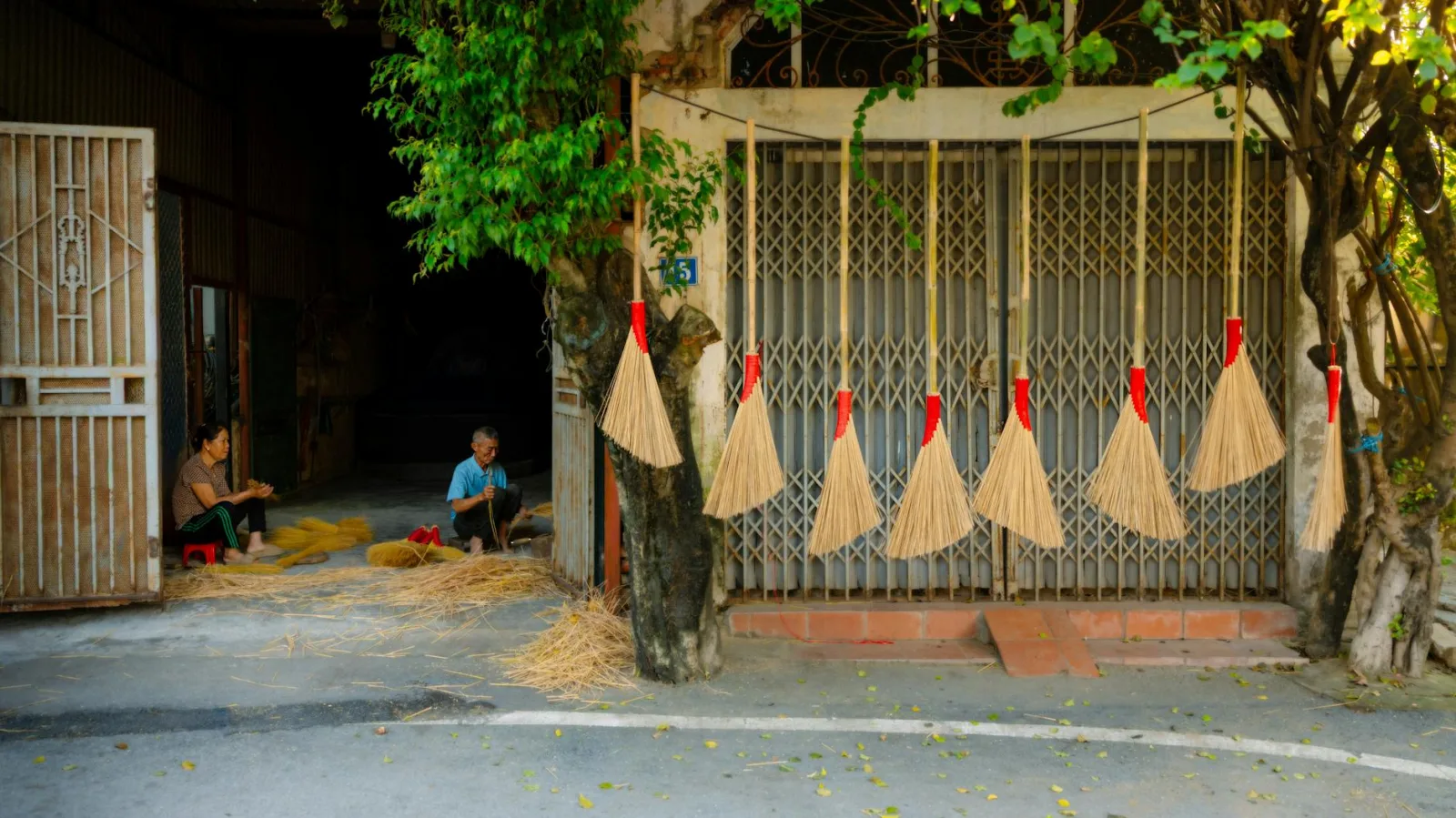 Two people making brooms outside a rural workshop with brooms hanging on the wall.