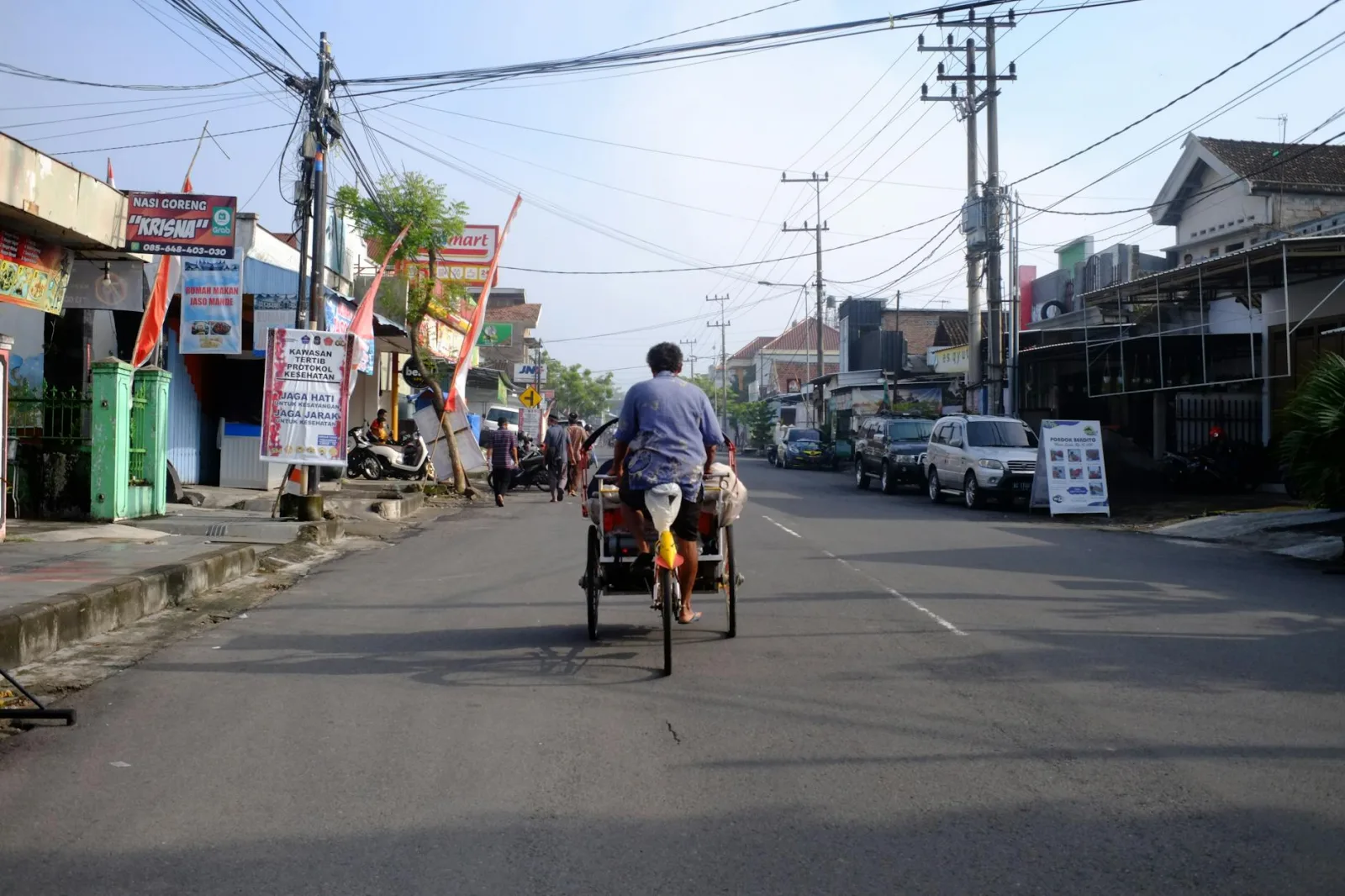 A becak rider traverses a busy street in Blitar, East Java, Indonesia under a clear sky, showcasing urban life.