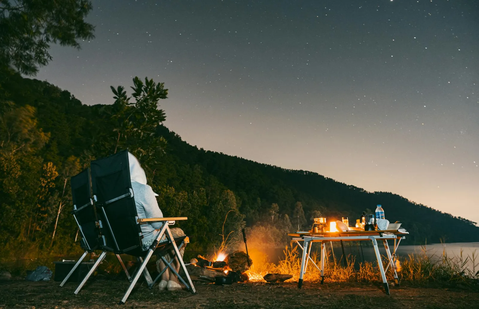 A serene camping scene by a lake with a campfire, chairs, and a star-filled sky.