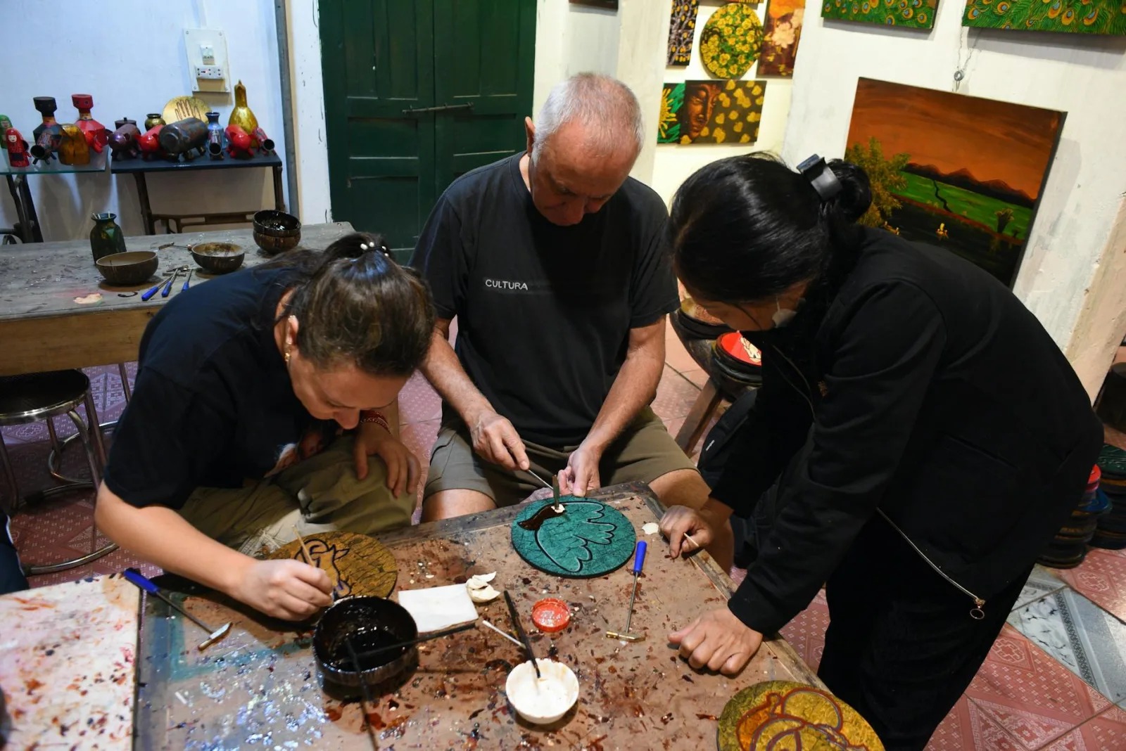 Three adults participating in a traditional Vietnamese art workshop, focused on creating lacquerware designs.