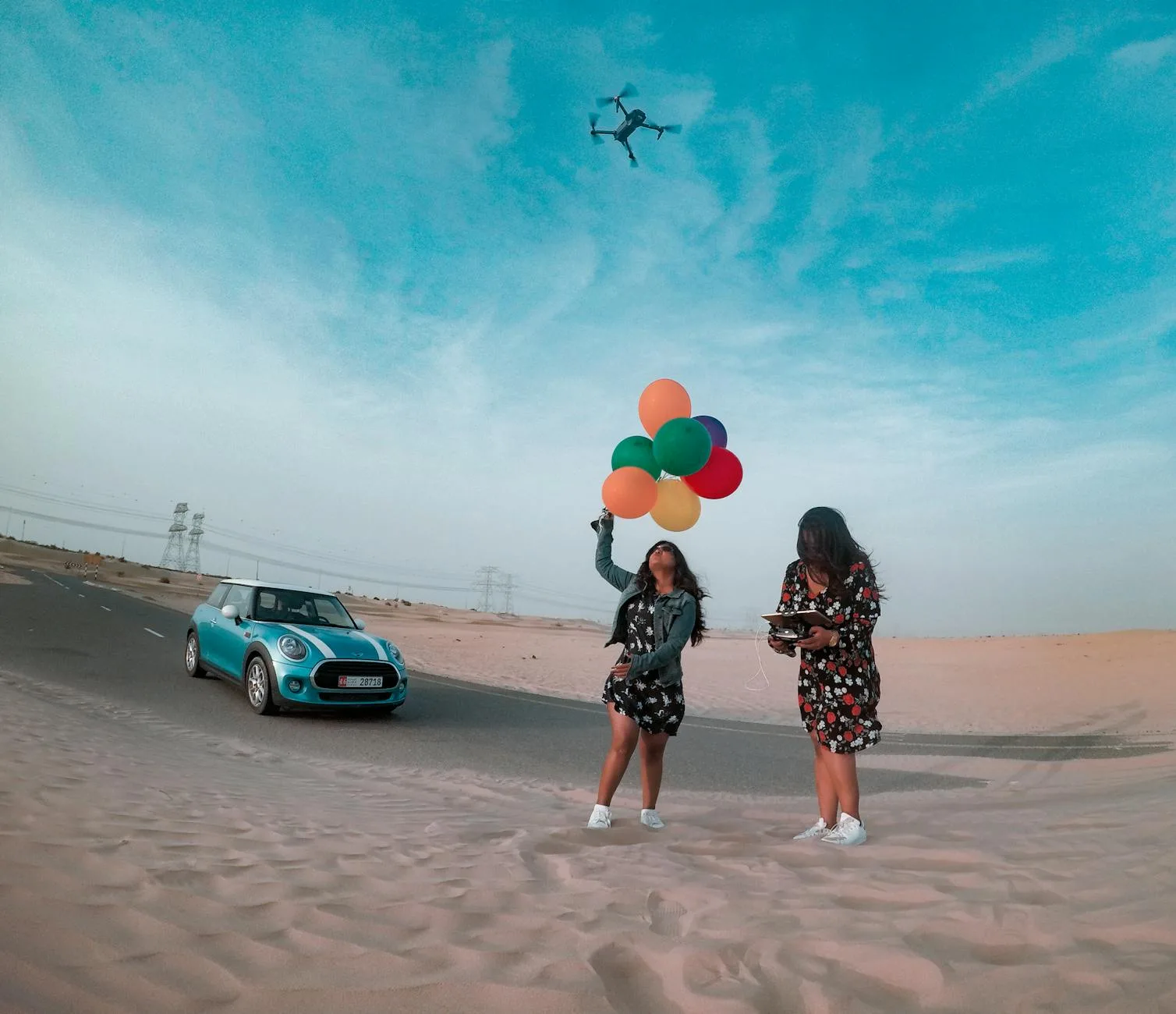 Two women with balloons and a Mini Cooper in the Dubai desert under a drone. Vibrant and joyous scene.