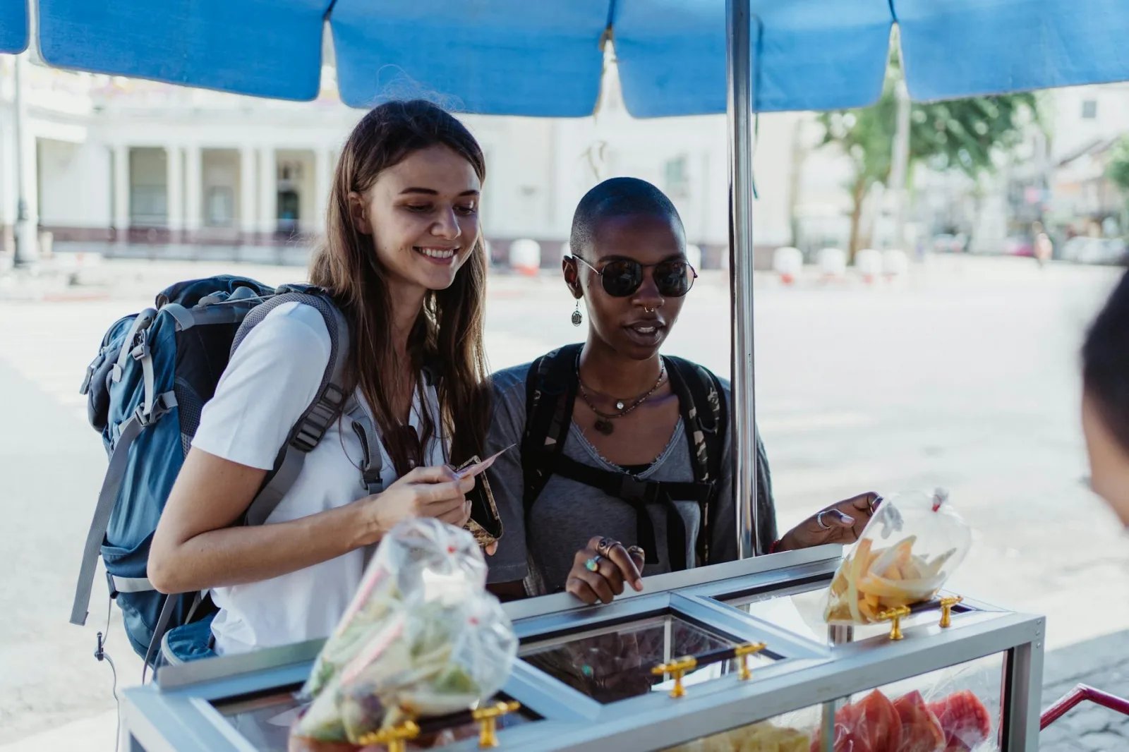Two women with backpacks exploring local street food in an urban outdoor setting.