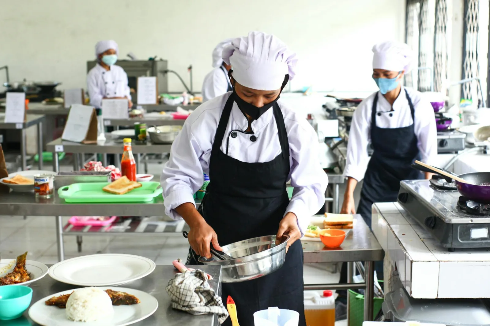 Chefs in masks preparing food in a commercial kitchen, emphasizing hygiene and teamwork.