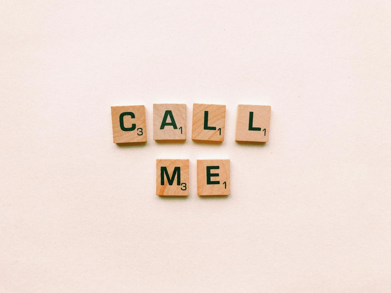 Wooden letter tiles spelling 'Call Me' on a pastel pink background, invoking communication and connection.