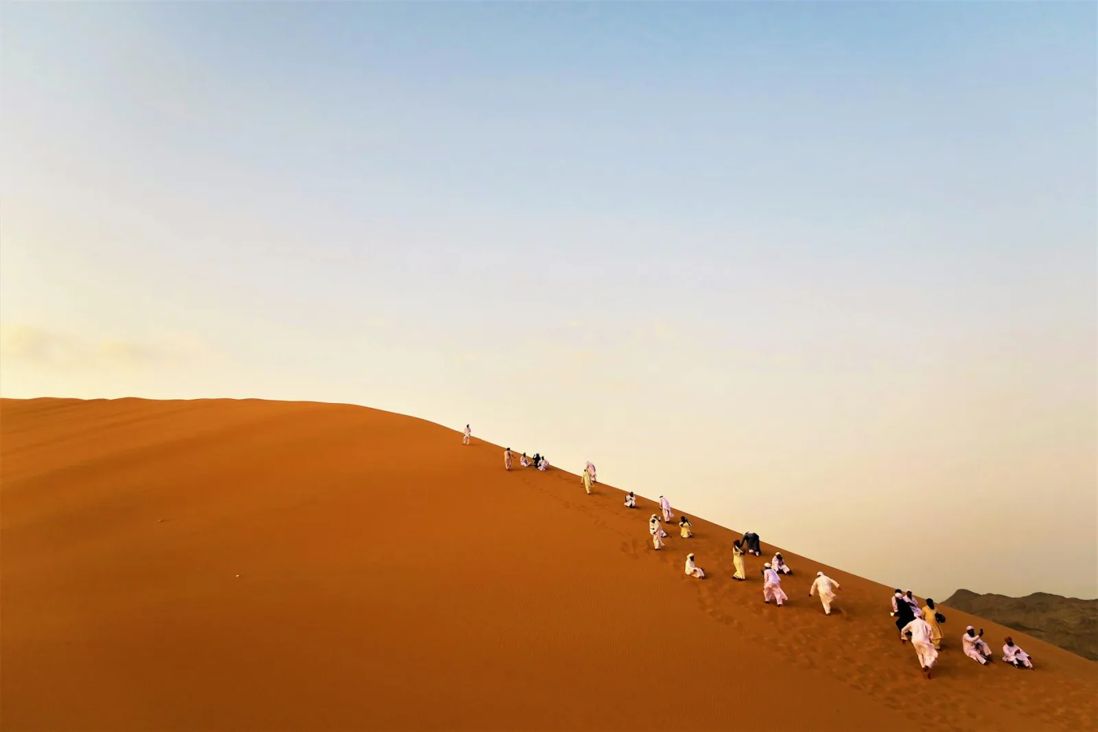 A group of adults climbs a large sand dune in the Bedir desert, Saudi Arabia under a clear sky.