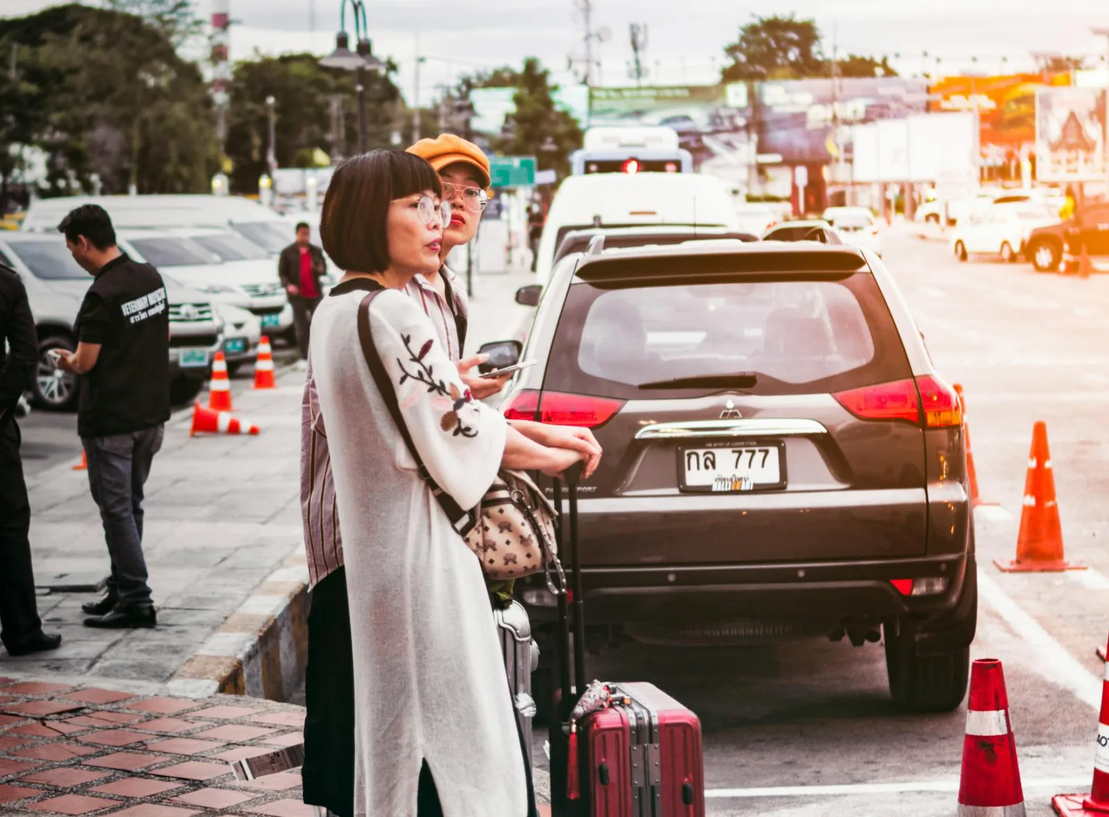 Two women with luggage waiting by a parked car on a busy street in Thailand.