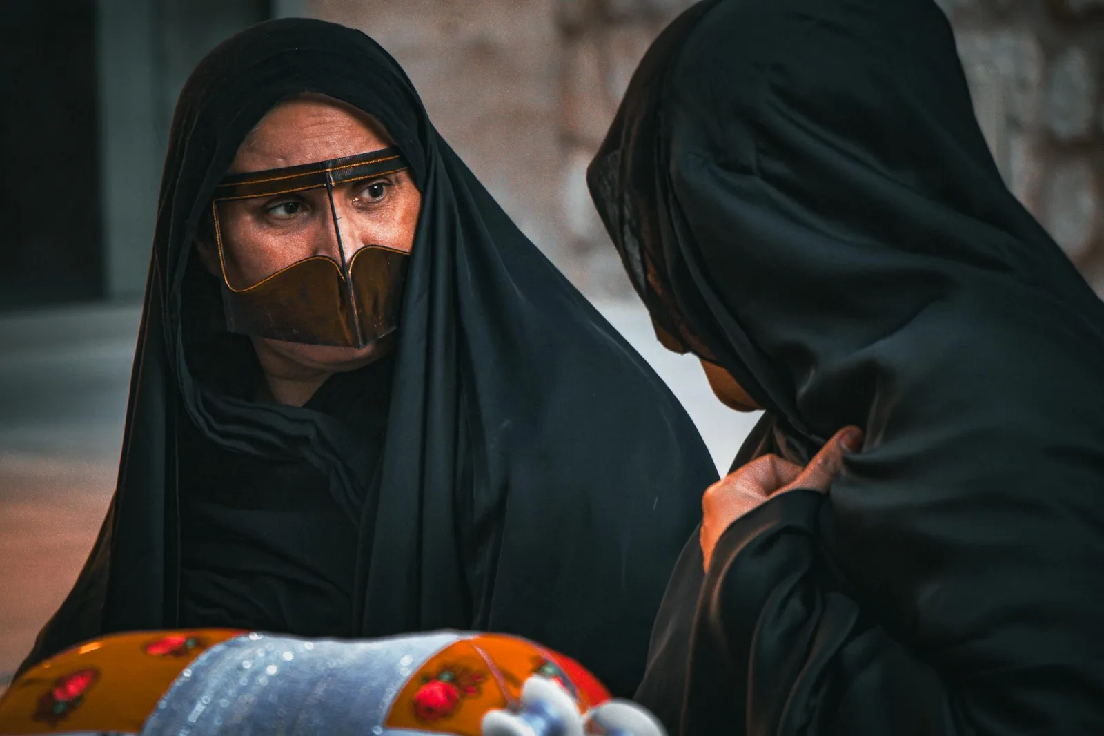 Two women in traditional Middle Eastern attire with brown face coverings and black abayas sharing a moment outdoors.