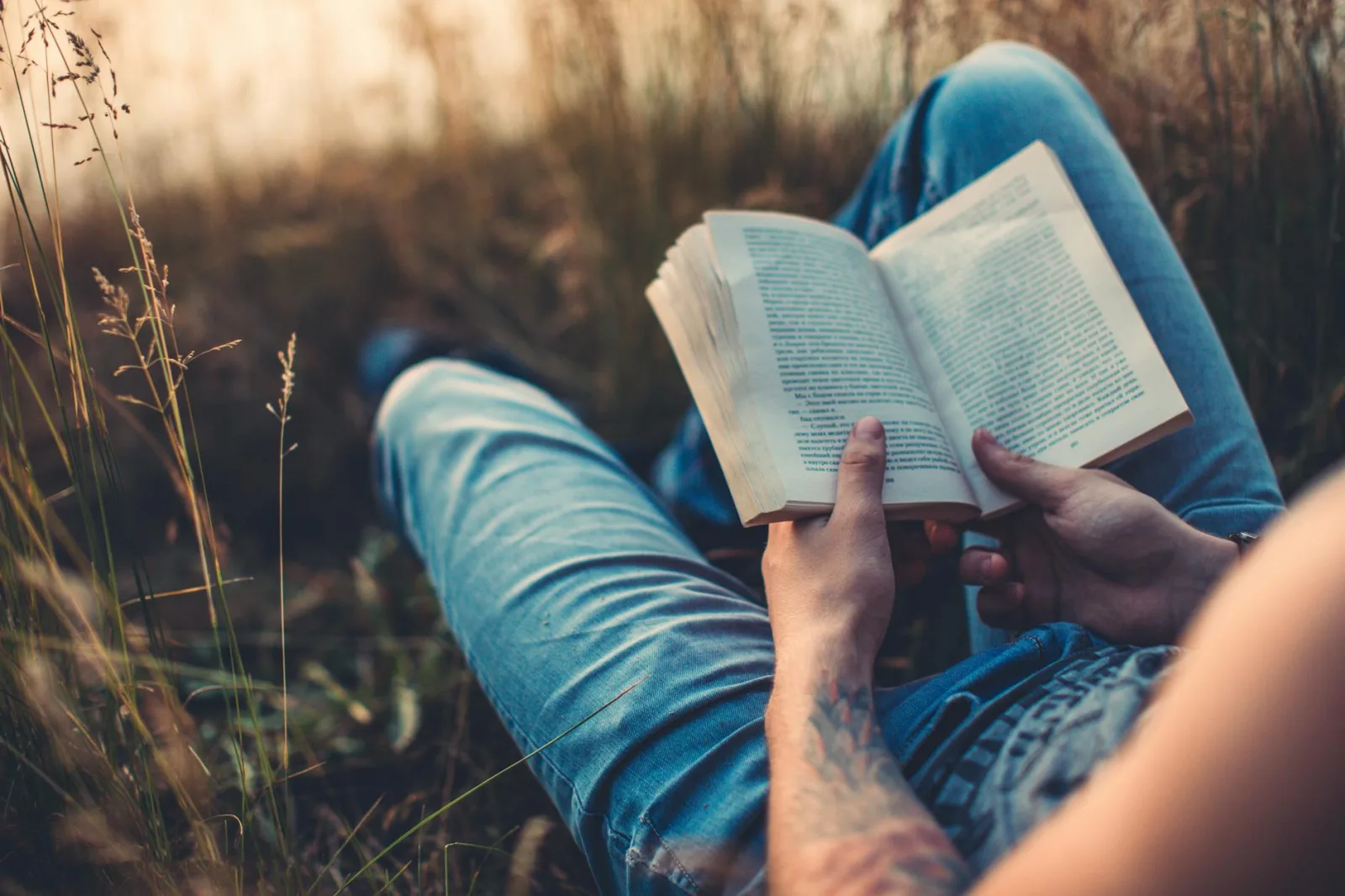 Person relaxing outdoors in denim jeans, reading a book on a sunny day.