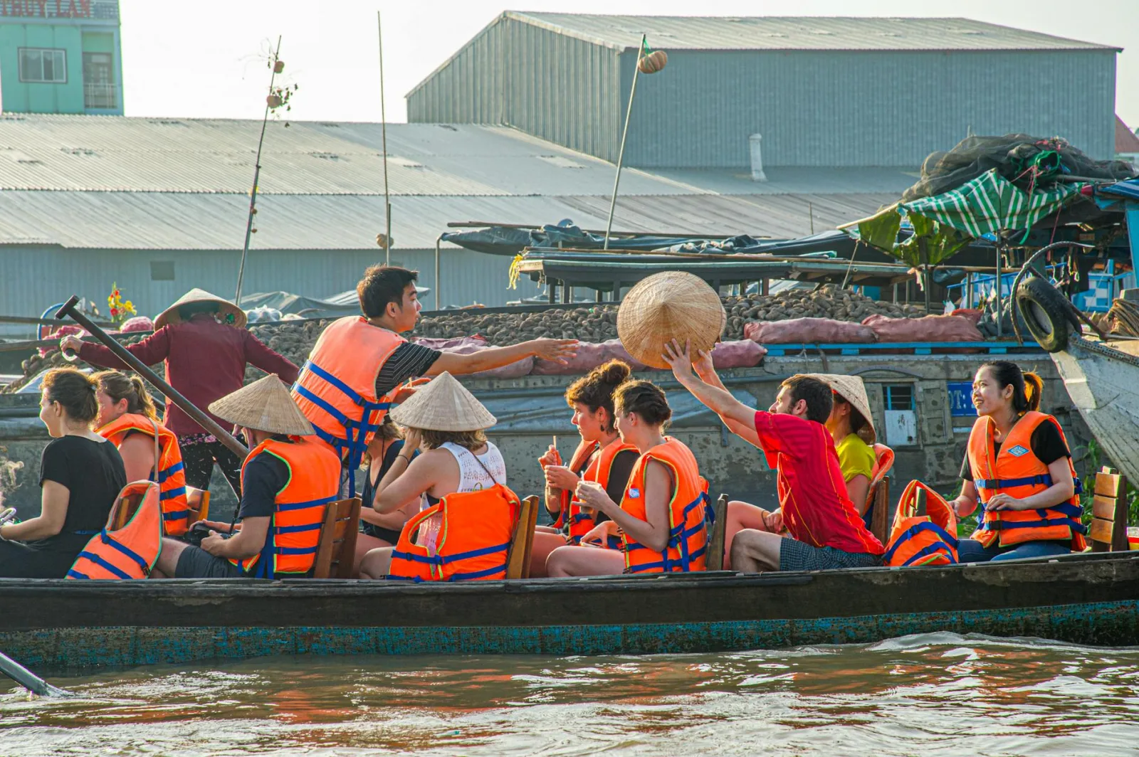 Tourists wearing life jackets and conical hats explore a vibrant floating market in a traditional boat.