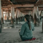 A woman meditates on sand beneath an ancient wooden pier by the sea.