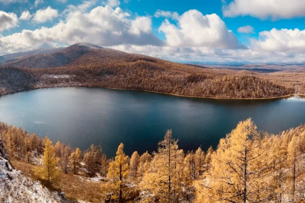 Tranquil lake surrounded by autumn forests and mountains under a bright blue sky.