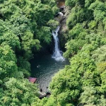 A picturesque aerial view of a waterfall surrounded by dense green forest and visible swimmers.