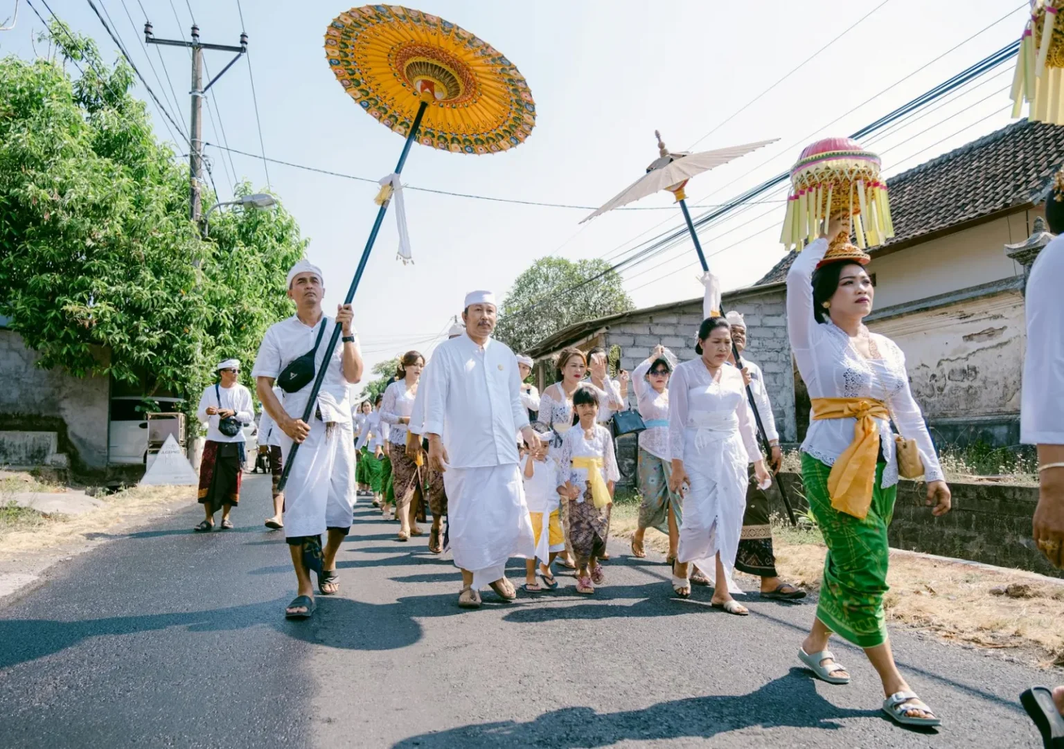A Balinese procession with traditional attire and vibrant umbrellas on a sunny day.